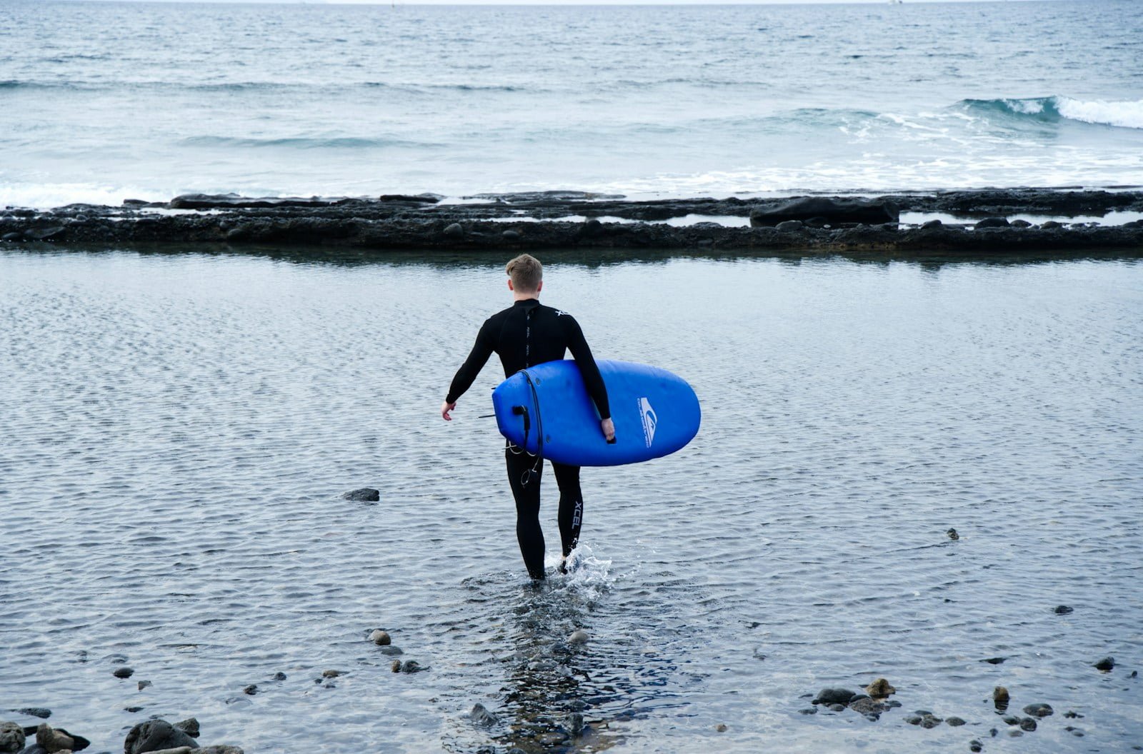 alquiler de equipos de surf en los cristianos - a man in a wet suit carrying a blue surfboard